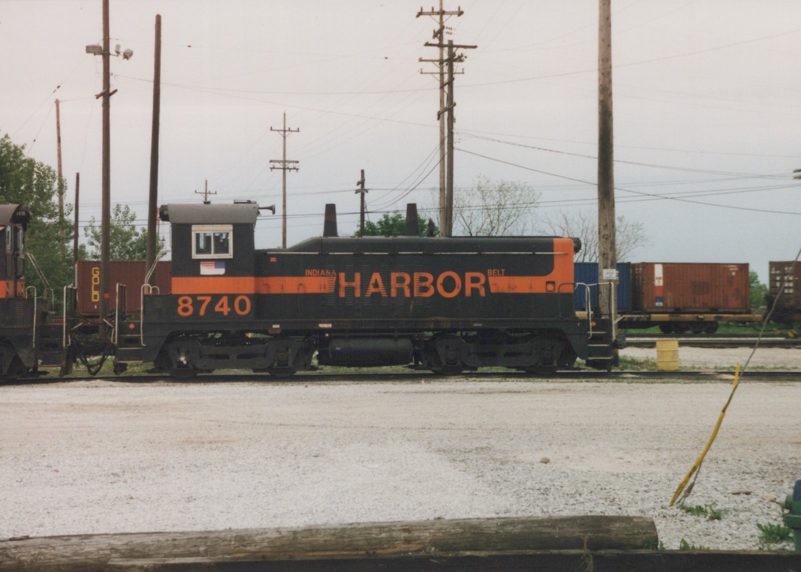 Side view of a black diesel switcher with an orange stripe, the number 8740 and the word HARBOR on its hood, coupled to another switcher, with container cars and utility poles behind it under an overcast sky.