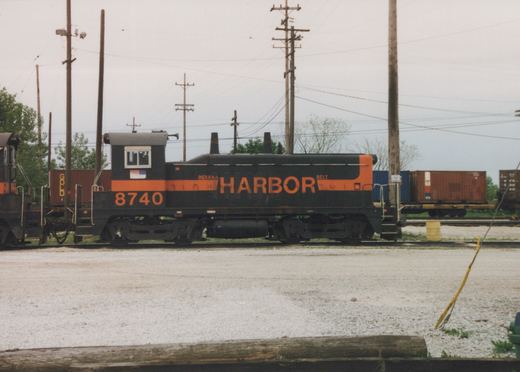 Side view of a black diesel switcher with an orange stripe, the number 8740 and the word HARBOR on its hood, coupled to another switcher, with container cars and utility poles behind it under an overcast sky.