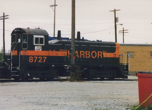A black diesel switcher with an orange stripe and the lettering INDIANA HARBOR BELT and 8727 stands on a track beside a gravel lot, with utility poles and a yellow brick building behind it.