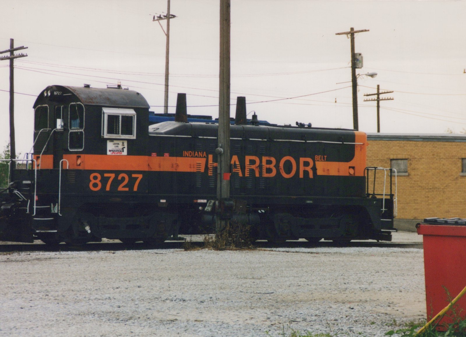 A black diesel switcher with an orange stripe and the lettering INDIANA HARBOR BELT and 8727 stands on a track beside a gravel lot, with utility poles and a yellow brick building behind it.