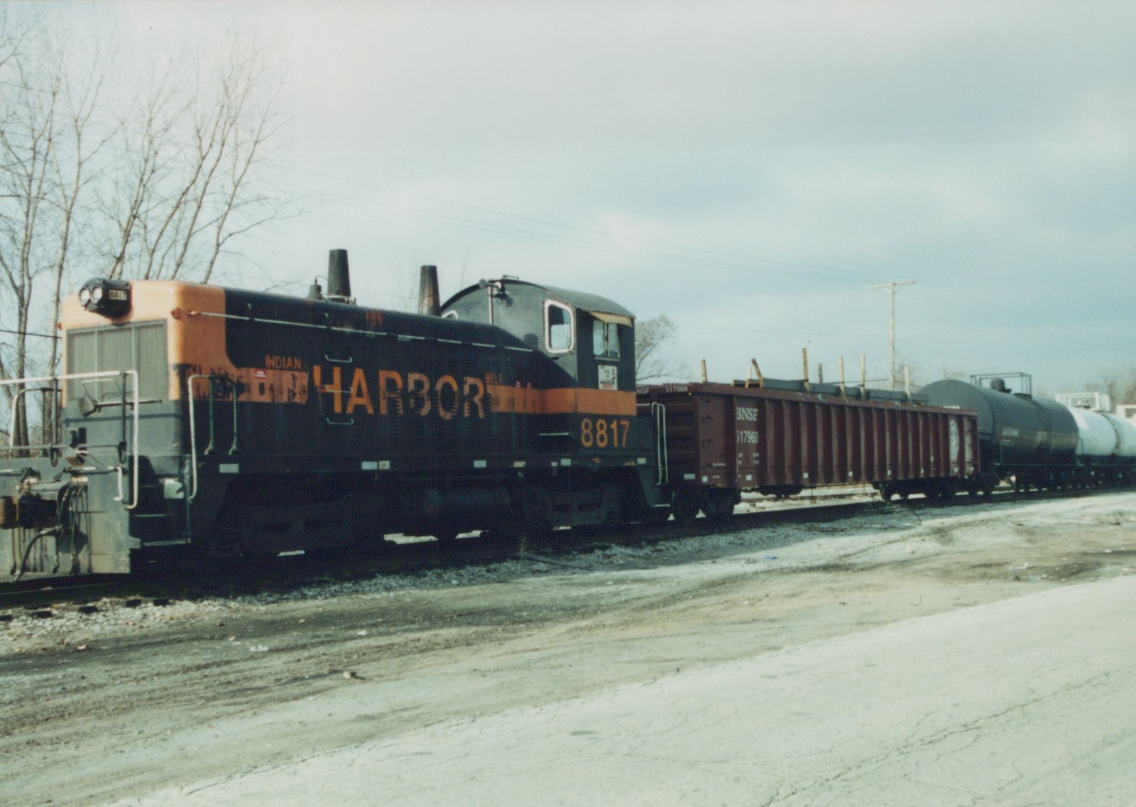 A black diesel switcher with a faded orange stripe, lettered HARBOR and numbered 8817, stands on a track beside a gravel lot. A brown BNSF gondola loaded with steel and a line of tank cars are coupled behind it, under an overcast sky with bare trees.