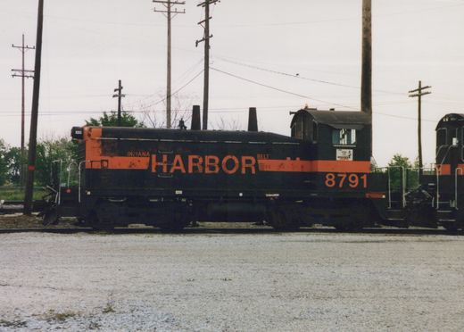 Side view of a weathered black diesel switcher with orange stripe and large orange HARBOR lettering, number 8791 on the cab, coupled to another black and orange switcher, beside a gravel area under an overcast sky.