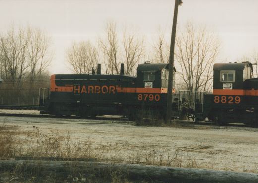 Side view of a black diesel switcher with an orange stripe, HARBOR lettered on its hood and 8790 on its cab, coupled near a second switcher numbered 8829, with bare trees and an overcast sky behind.