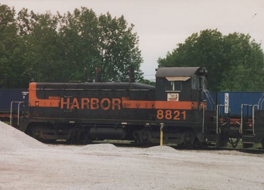 Side view of a black diesel switcher with an orange stripe and INDIANA HARBOR BELT lettering, numbered 8821, beside piles of gravel with blue containers and trees behind it.