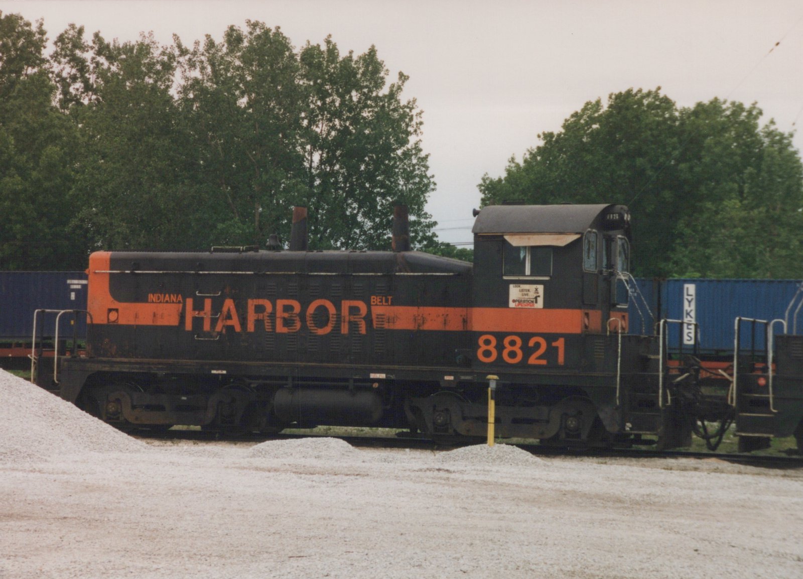 Side view of a black diesel switcher with an orange stripe and INDIANA HARBOR BELT lettering, numbered 8821, beside piles of gravel with blue containers and trees behind it.