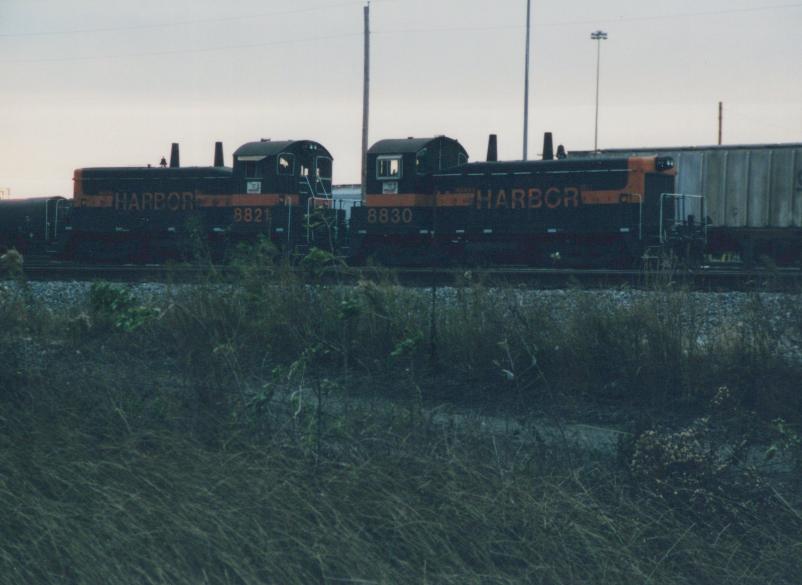 Two black and orange diesel switchers lettered HARBOR, numbered 8821 and 8830, stand cab to cab on a yard track behind a weedy embankment, with a gray covered hopper on the right and a tank car on the left.
