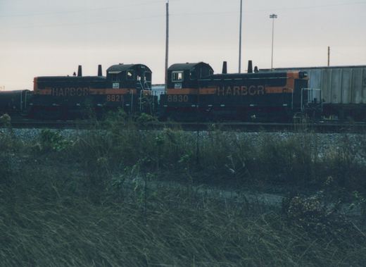 Two black and orange diesel switchers lettered HARBOR, numbered 8821 and 8830, stand cab to cab on a yard track behind a weedy embankment, with a gray covered hopper on the right and a tank car on the left.