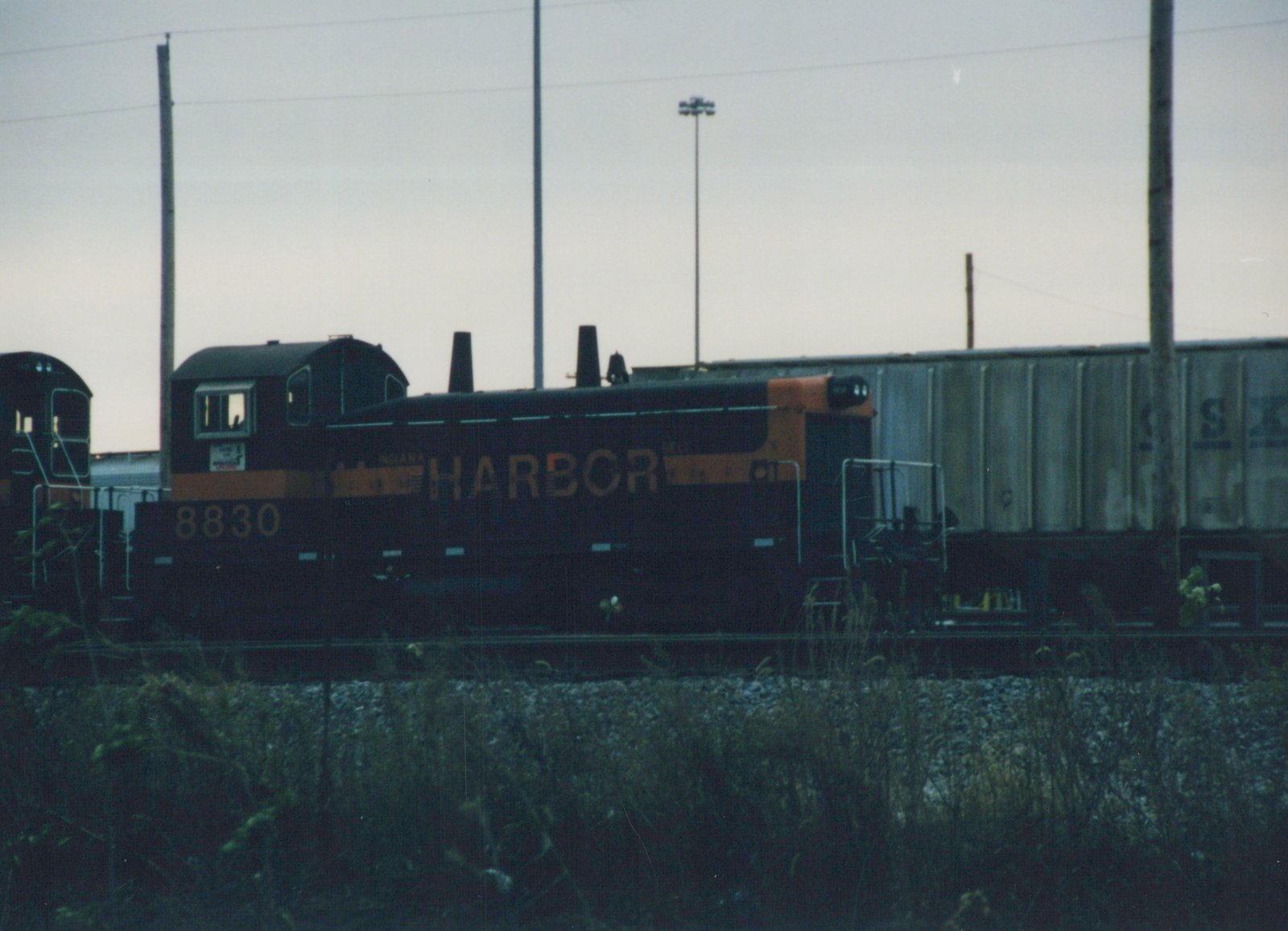 Side view of a black-and-orange end-cab diesel switcher numbered 8830 and lettered Harbor, standing on a yard track behind tall weeds, with a grey covered hopper and another switcher's cab nearby under an overcast evening sky.