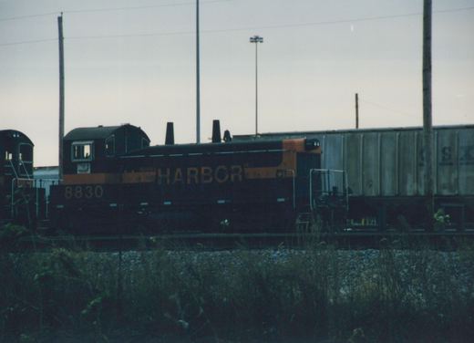 Side view of a black-and-orange end-cab diesel switcher numbered 8830 and lettered Harbor, standing on a yard track behind tall weeds, with a grey covered hopper and another switcher's cab nearby under an overcast evening sky.