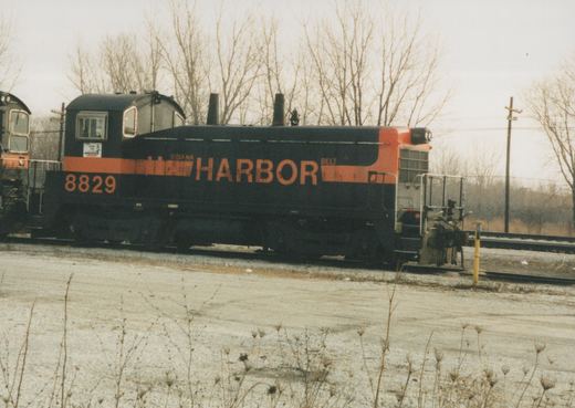 Side view of a black diesel switcher with an orange stripe, lettered HARBOR and numbered 8829, coupled to another locomotive on a yard track under bare winter trees.