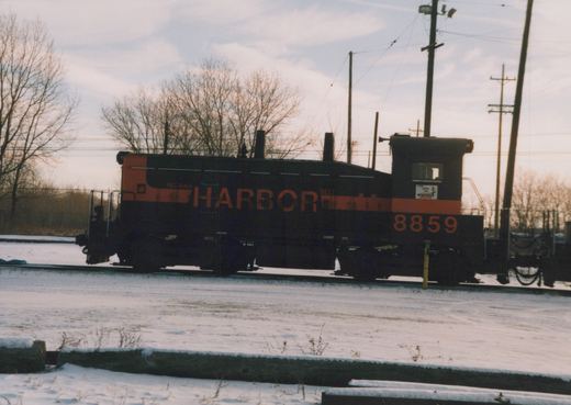 Side view of a black and orange end-cab diesel switcher lettered INDIANA HARBOR BELT and numbered 8859, standing on a track in snow under bare trees and utility poles.