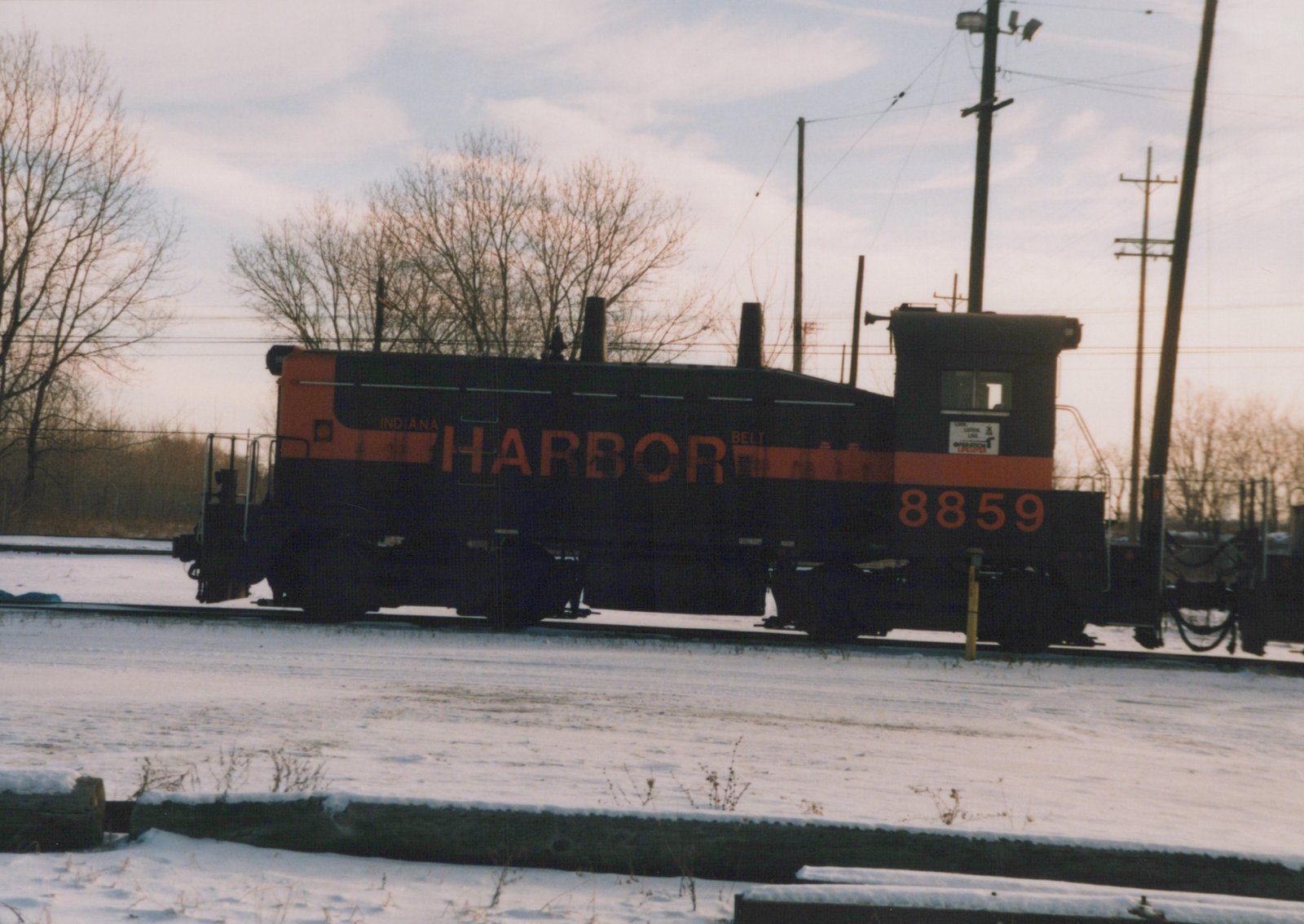 Side view of a black and orange end-cab diesel switcher lettered INDIANA HARBOR BELT and numbered 8859, standing on a track in snow under bare trees and utility poles.
