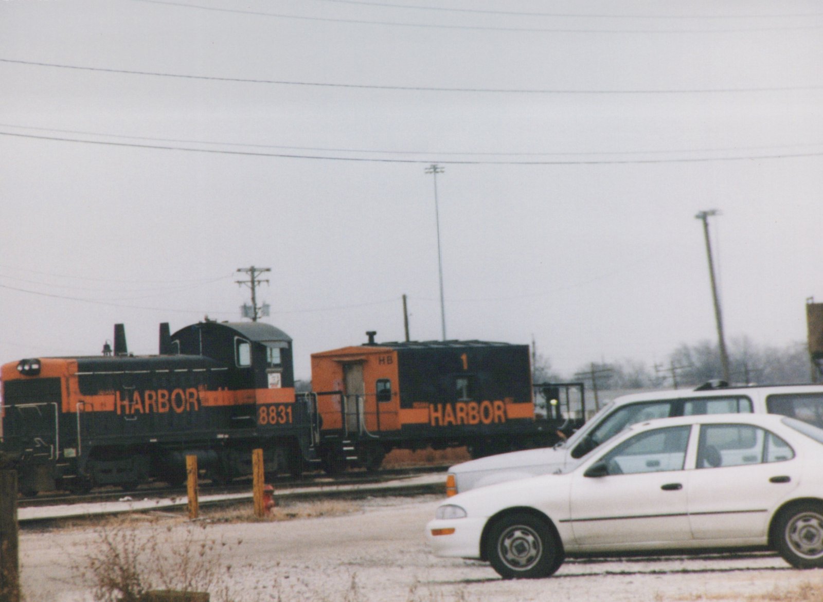 A black and orange end-cab switcher lettered HARBOR, number 8831, coupled to a short black and orange caboose numbered 1, on yard tracks behind two parked white cars on an overcast winter day.