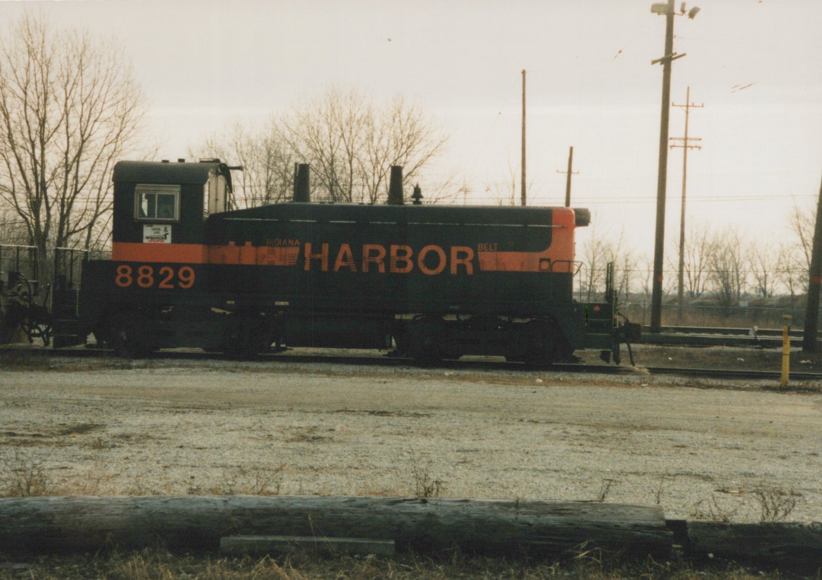 A black diesel switcher with an orange stripe and the large orange word HARBOR, numbered 8829 on the cab, sits on yard tracks behind a gravel lot under a gray sky with bare trees.