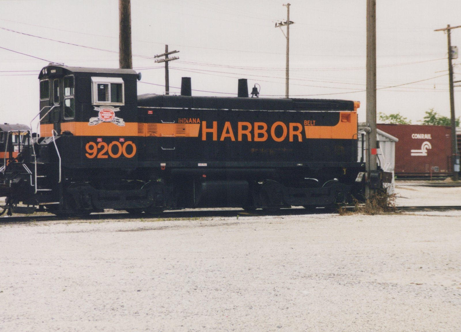 Side view of a black diesel switcher with an orange stripe and large orange HARBOR lettering, number 9200, standing on gravel yard track under an overcast sky.