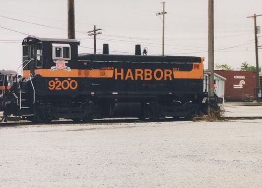 Side view of a black diesel switcher with an orange stripe and large orange HARBOR lettering, number 9200, standing on gravel yard track under an overcast sky.
