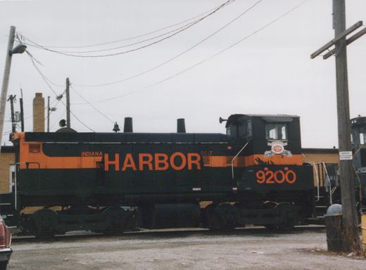 Side view of a black switcher locomotive with an orange stripe, HARBOR in large orange letters on the hood and 9200 on the cab, standing in front of brick buildings under utility wires.