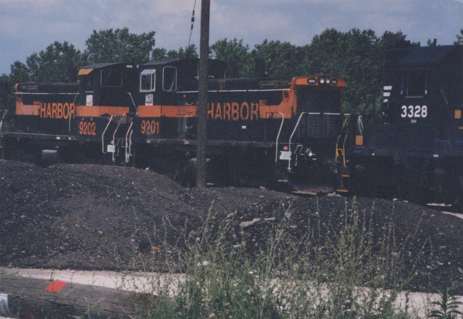 Two black diesel switchers with orange stripes and HARBOR lettering stand behind a pile of ballast next to a wooden pole, with a black locomotive numbered 3328 at the right.