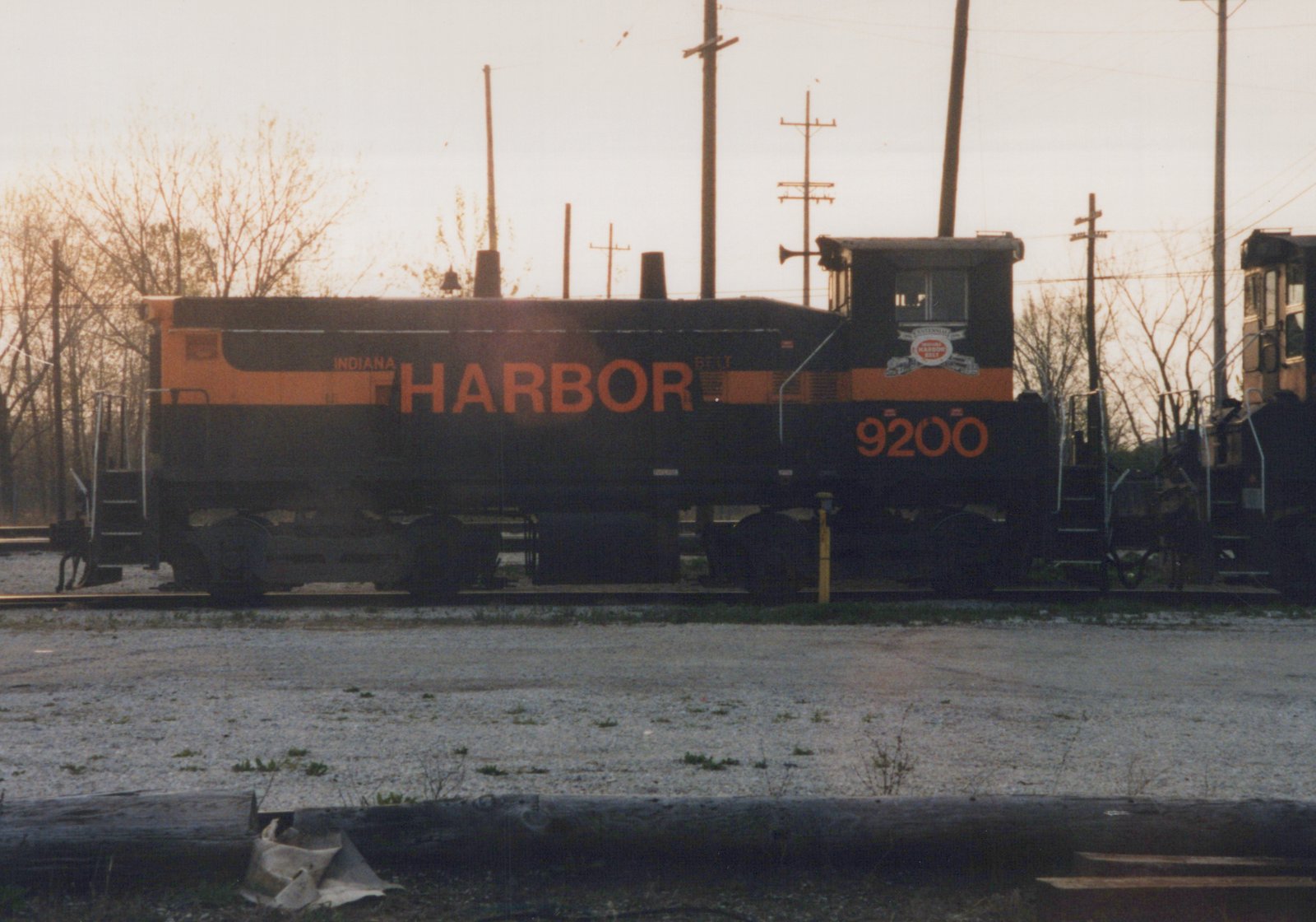Side view of a black and orange diesel switcher lettered INDIANA HARBOR BELT, number 9200, standing on a gravel yard track with bare trees and utility poles behind it under a pale sky.