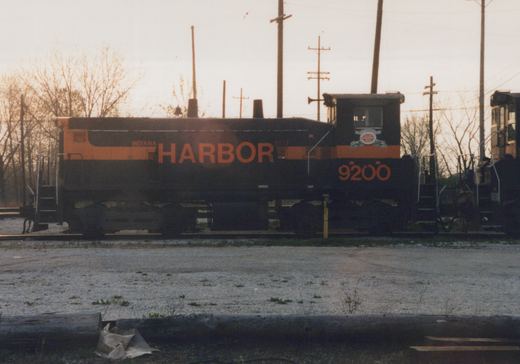 Side view of a black and orange diesel switcher lettered INDIANA HARBOR BELT, number 9200, standing on a gravel yard track with bare trees and utility poles behind it under a pale sky.