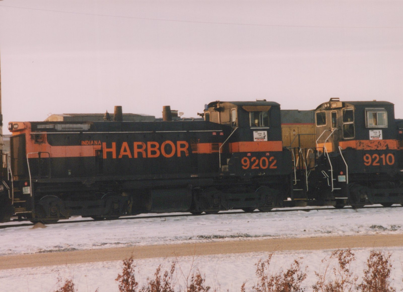 Side view of two black and orange Indiana Harbor Belt switcher locomotives, numbered 9202 and 9210, with a yellow locomotive between them on a snowy track.