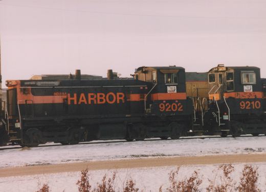 Side view of two black and orange Indiana Harbor Belt switcher locomotives, numbered 9202 and 9210, with a yellow locomotive between them on a snowy track.