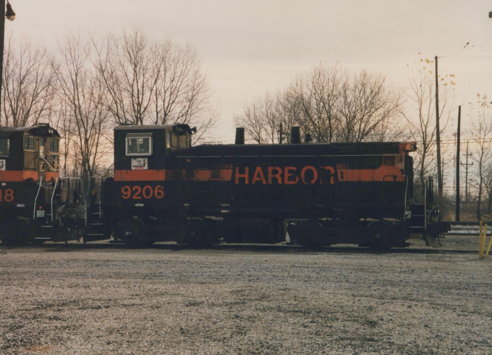 A black diesel switcher with an orange stripe, the number 9206 on its cab and HARBOR in large orange letters on its hood, stands on a gravel-covered track in front of bare trees and a gray sky, with part of a second black and orange locomotive at the left.