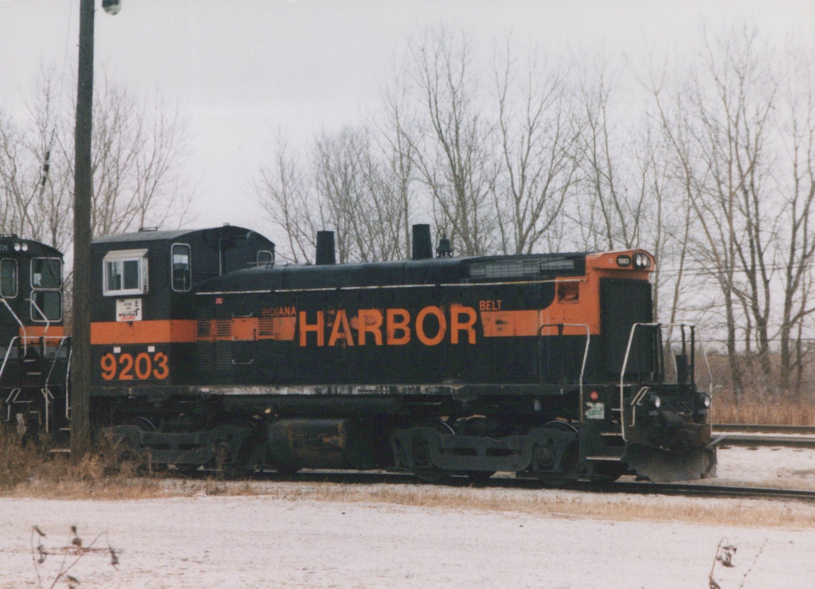 A black diesel switcher with an orange stripe and large orange HARBOR lettering, number 9203 on the cab, stands on track in a light snowfall in front of bare trees.