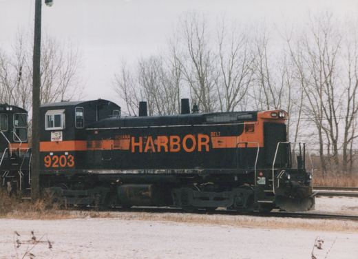A black diesel switcher with an orange stripe and large orange HARBOR lettering, number 9203 on the cab, stands on track in a light snowfall in front of bare trees.