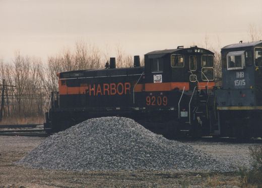 A black diesel switcher with an orange stripe and HARBOR in large orange letters, numbered 9209, stands behind a pile of crushed stone, next to the cab of a blue-gray switcher stenciled IHB 1505, under an overcast sky with bare trees.