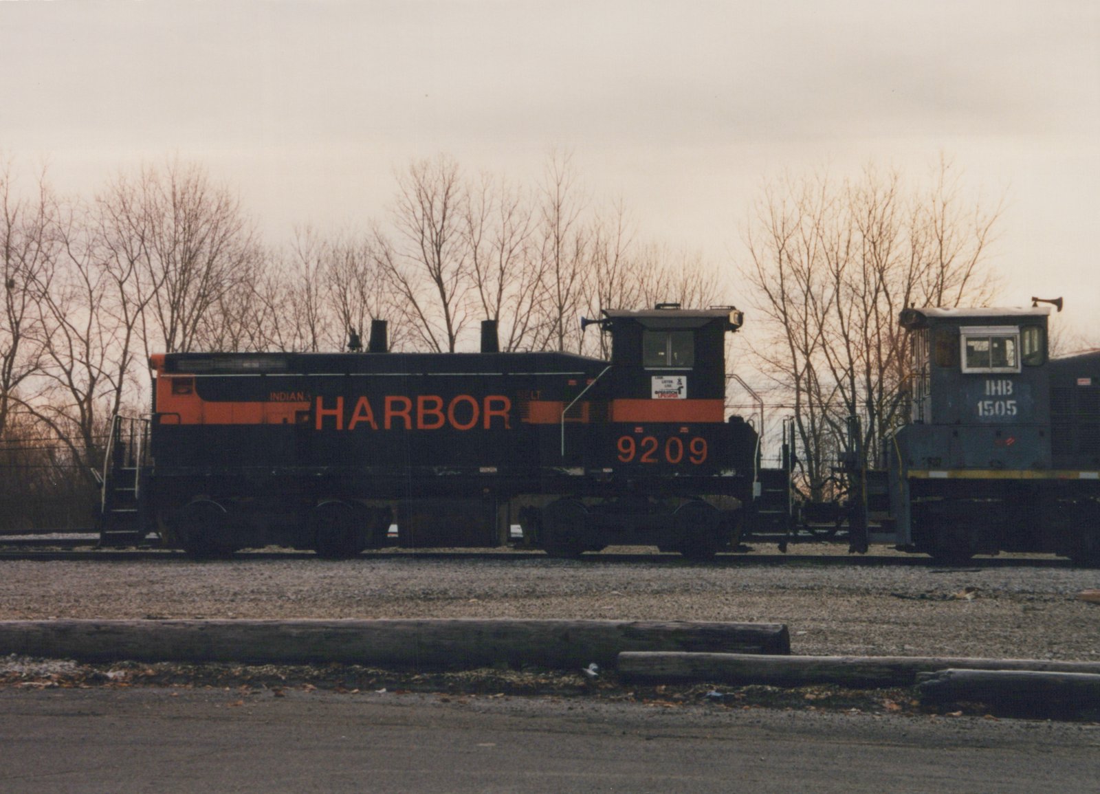 A black diesel switcher with an orange stripe and HARBOR in large orange letters stands on a yard track next to a grey switcher, with bare trees under an overcast sky behind them.