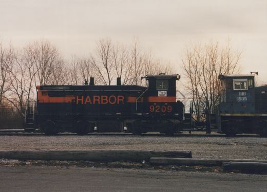 A black diesel switcher with an orange stripe and HARBOR in large orange letters stands on a yard track next to a grey switcher, with bare trees under an overcast sky behind them.