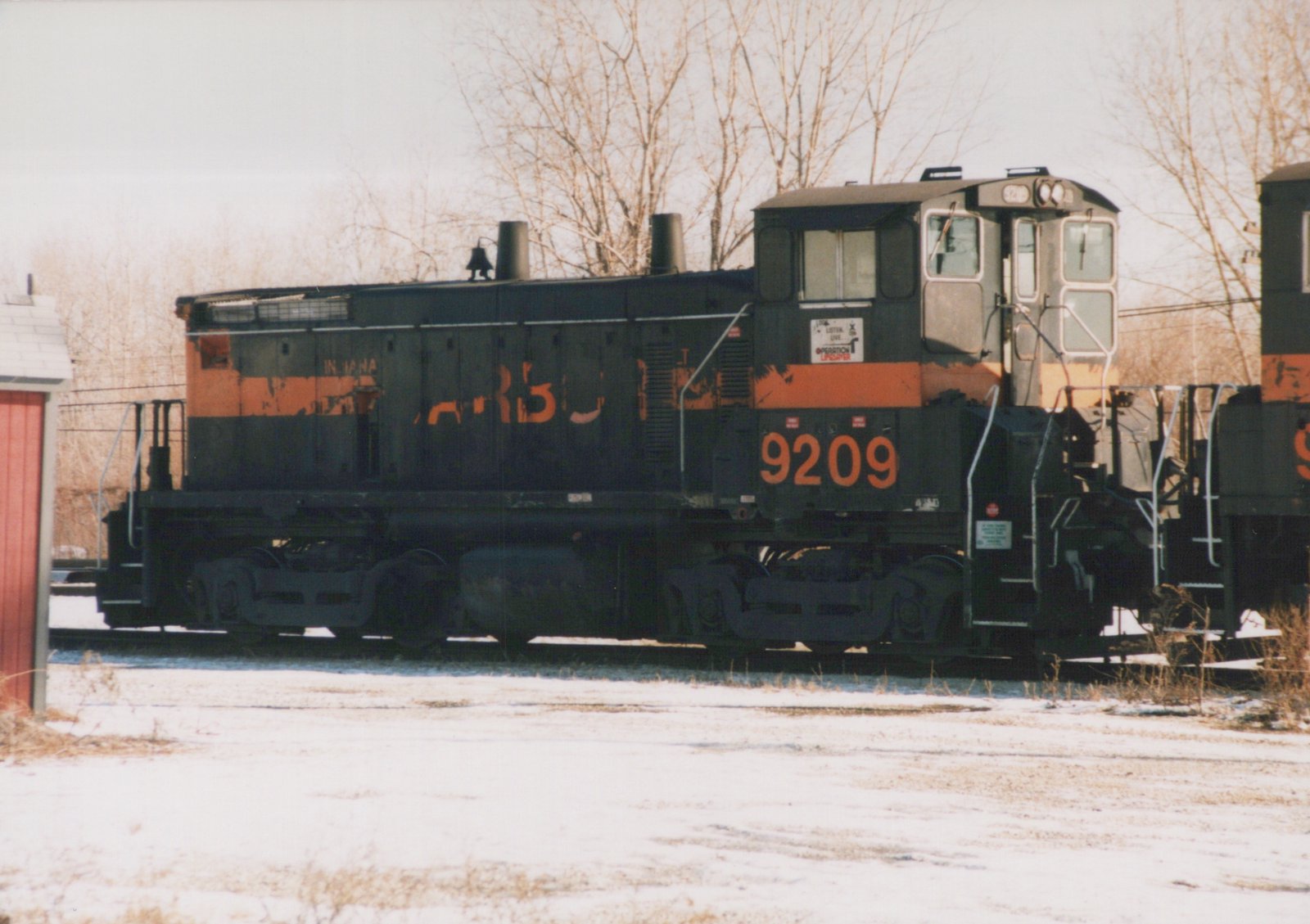 A black switcher locomotive with a faded orange stripe and the number 9209 on its cab, coupled to another locomotive, on snowy ground with bare trees behind.