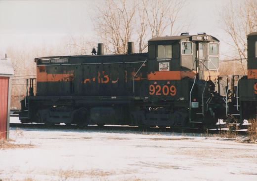 A black switcher locomotive with a faded orange stripe and the number 9209 on its cab, coupled to another locomotive, on snowy ground with bare trees behind.