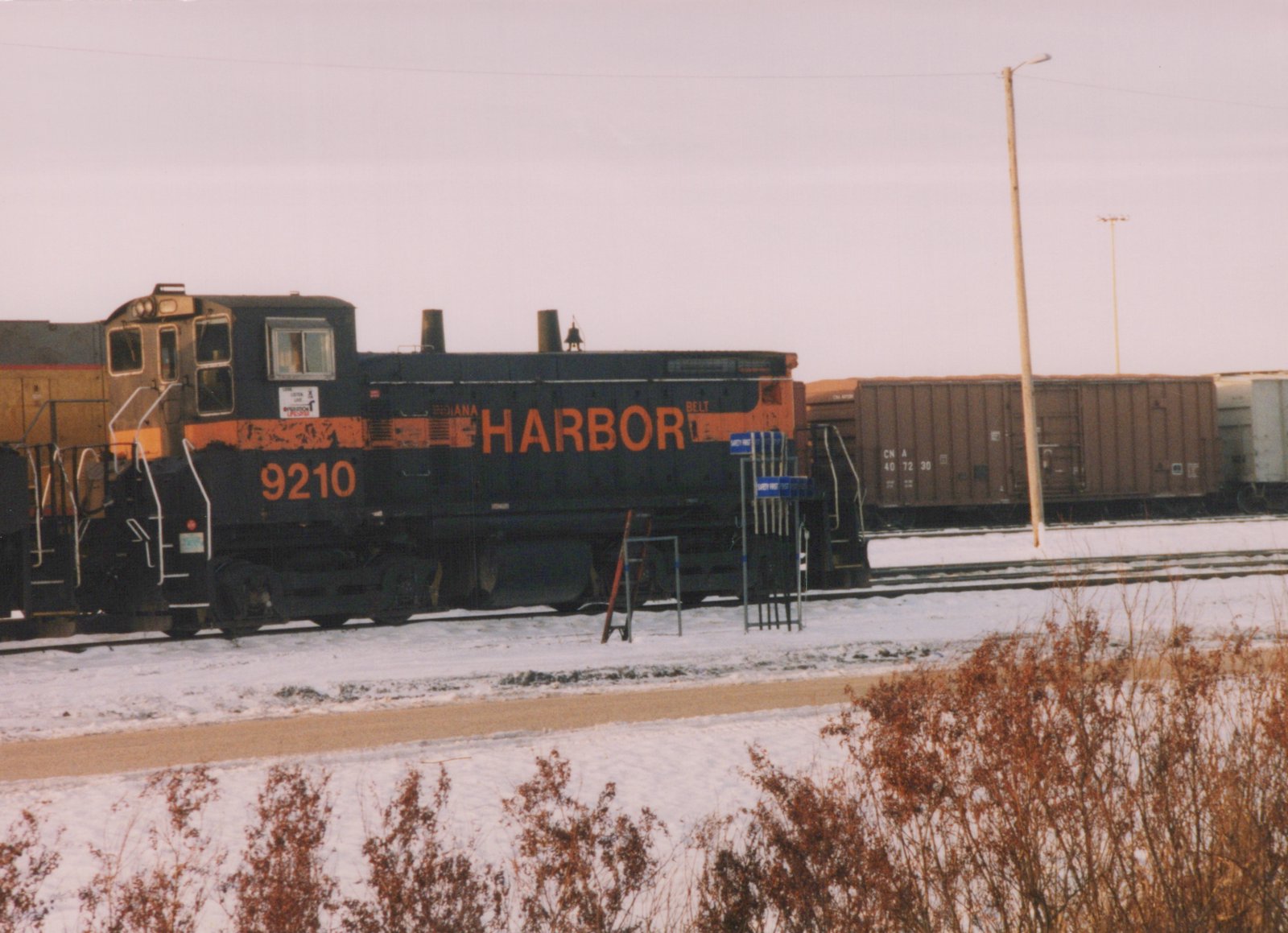 A black end-cab switcher with orange HARBOR lettering and the number 9210 stands on a snowy yard track, with a brown Canadian National boxcar behind it and dry brown bushes in the foreground.
