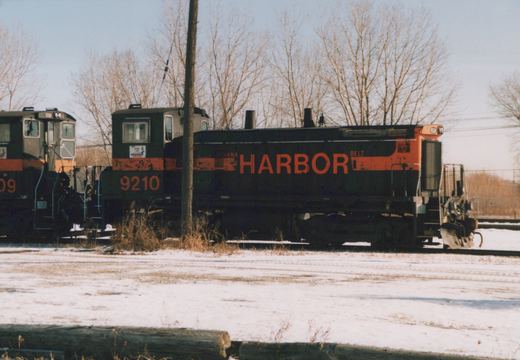 A black and orange diesel switcher lettered HARBOR with number 9210 on its cab, coupled cab to cab with another switcher, stands on snowy ground behind a utility pole under bare trees.
