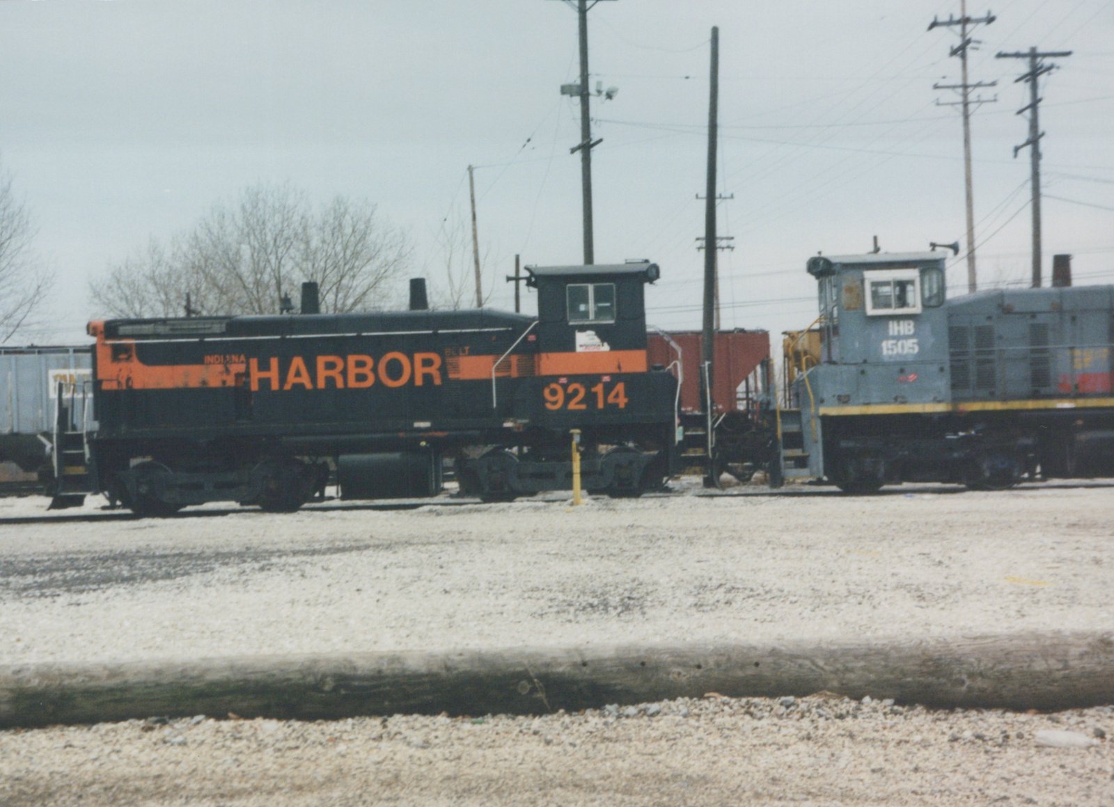 A black diesel switcher with an orange stripe and HARBOR lettering, numbered 9214, stands in a gravel yard beside a gray switcher numbered 1505, under an overcast sky with bare trees and utility poles.