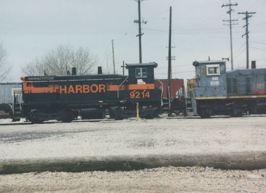 A black diesel switcher with an orange stripe and HARBOR lettering, numbered 9214, stands in a gravel yard beside a gray switcher numbered 1505, under an overcast sky with bare trees and utility poles.