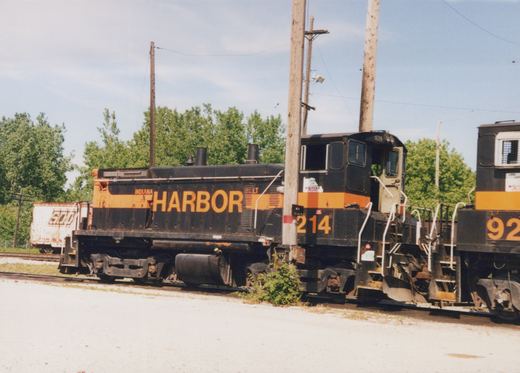 A weathered black switcher locomotive with an orange stripe and HARBOR in large orange letters stands on a yard track beside a gravel lot, coupled to a second black and orange locomotive, under utility poles and summer trees.