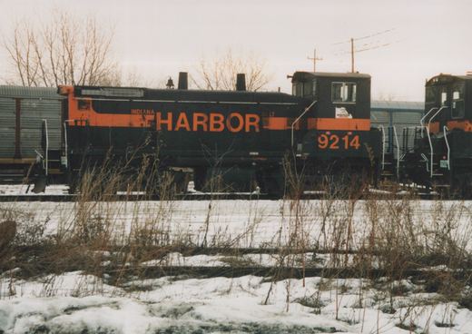 Side view of a black diesel switcher with an orange stripe, lettered HARBOR and numbered 9214, on a track beyond snowy ground and dry weeds, under an overcast sky.