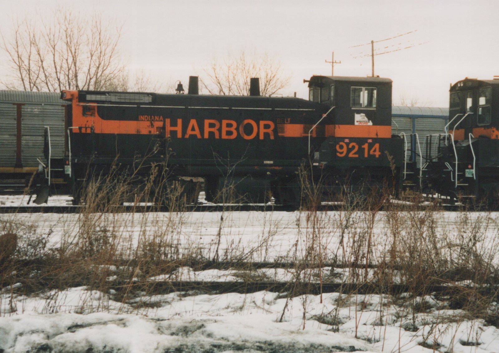 Side view of a black diesel switcher with an orange stripe, lettered HARBOR and numbered 9214, on a track beyond snowy ground and dry weeds, under an overcast sky.