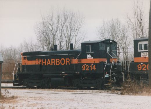A black diesel switcher with an orange stripe and large orange HARBOR lettering and the number 9214 stands on a track beside snow-dusted ground and bare trees, with the cab of a second black and orange locomotive at the right edge.
