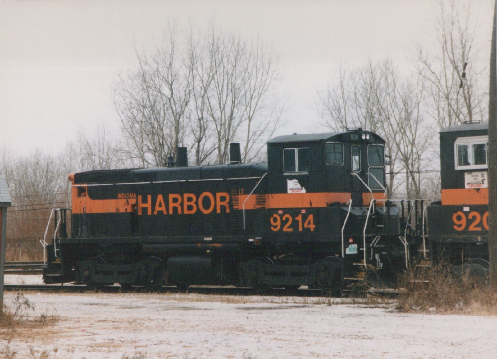 A black diesel switcher with an orange stripe and large orange HARBOR lettering and the number 9214 stands on a track beside snow-dusted ground and bare trees, with the cab of a second black and orange locomotive at the right edge.