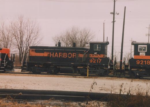 A black diesel switcher with orange stripes and INDIANA HARBOR lettering, numbered 9217, stands on a yard track beside a second unit numbered 9218, with bare trees and utility poles under a gray sky.