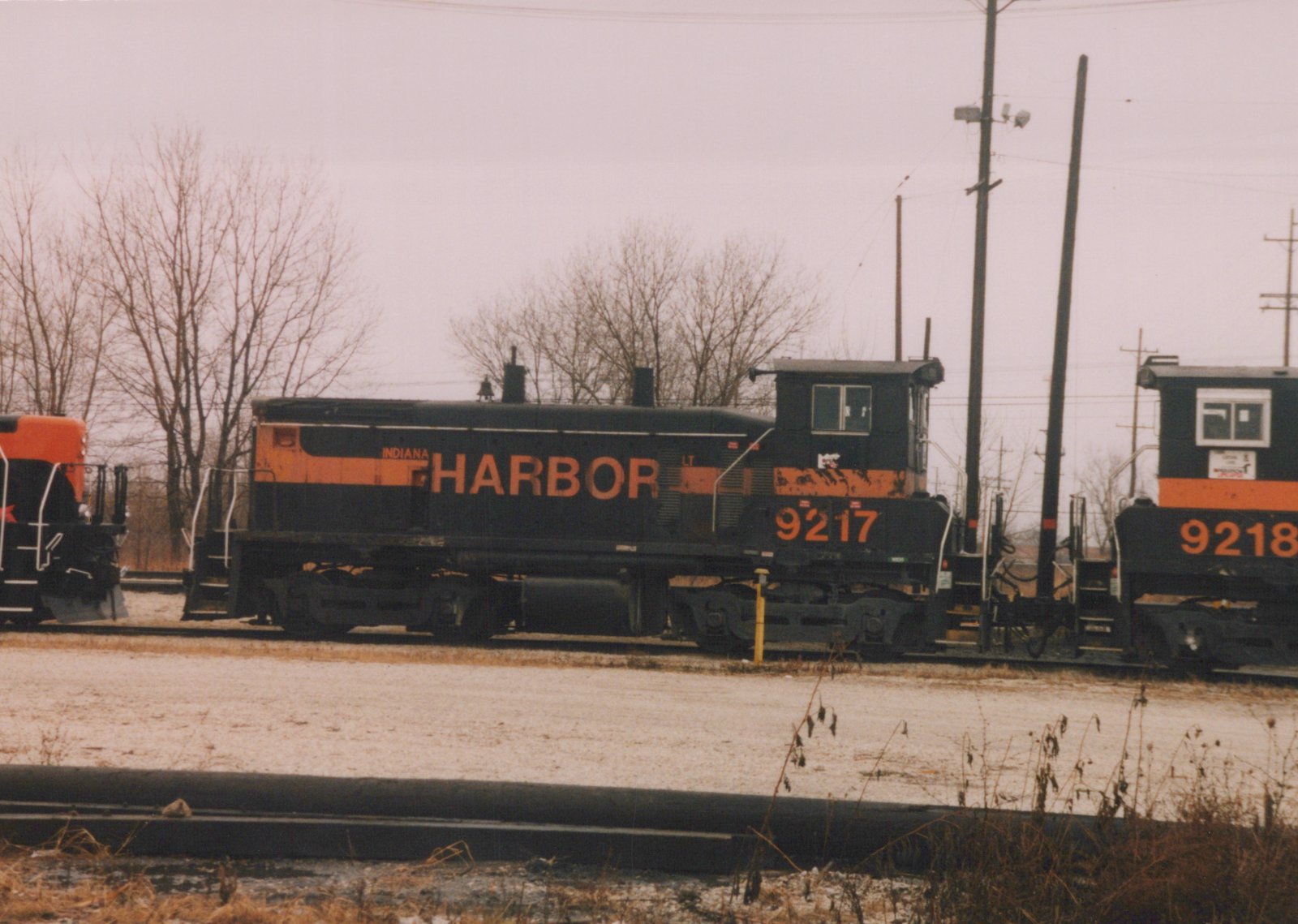 A black diesel switcher with orange stripes and INDIANA HARBOR lettering, numbered 9217, stands on a yard track beside a second unit numbered 9218, with bare trees and utility poles under a gray sky.