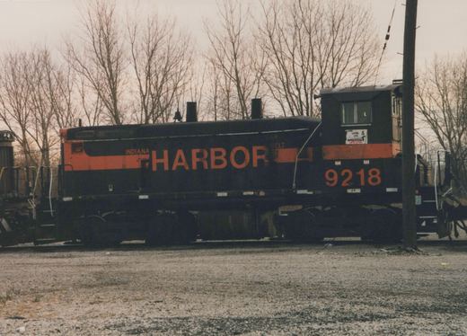 A black diesel switcher with an orange stripe and large orange HARBOR lettering and the number 9218 stands on a gravel yard track in front of bare trees under an overcast sky.