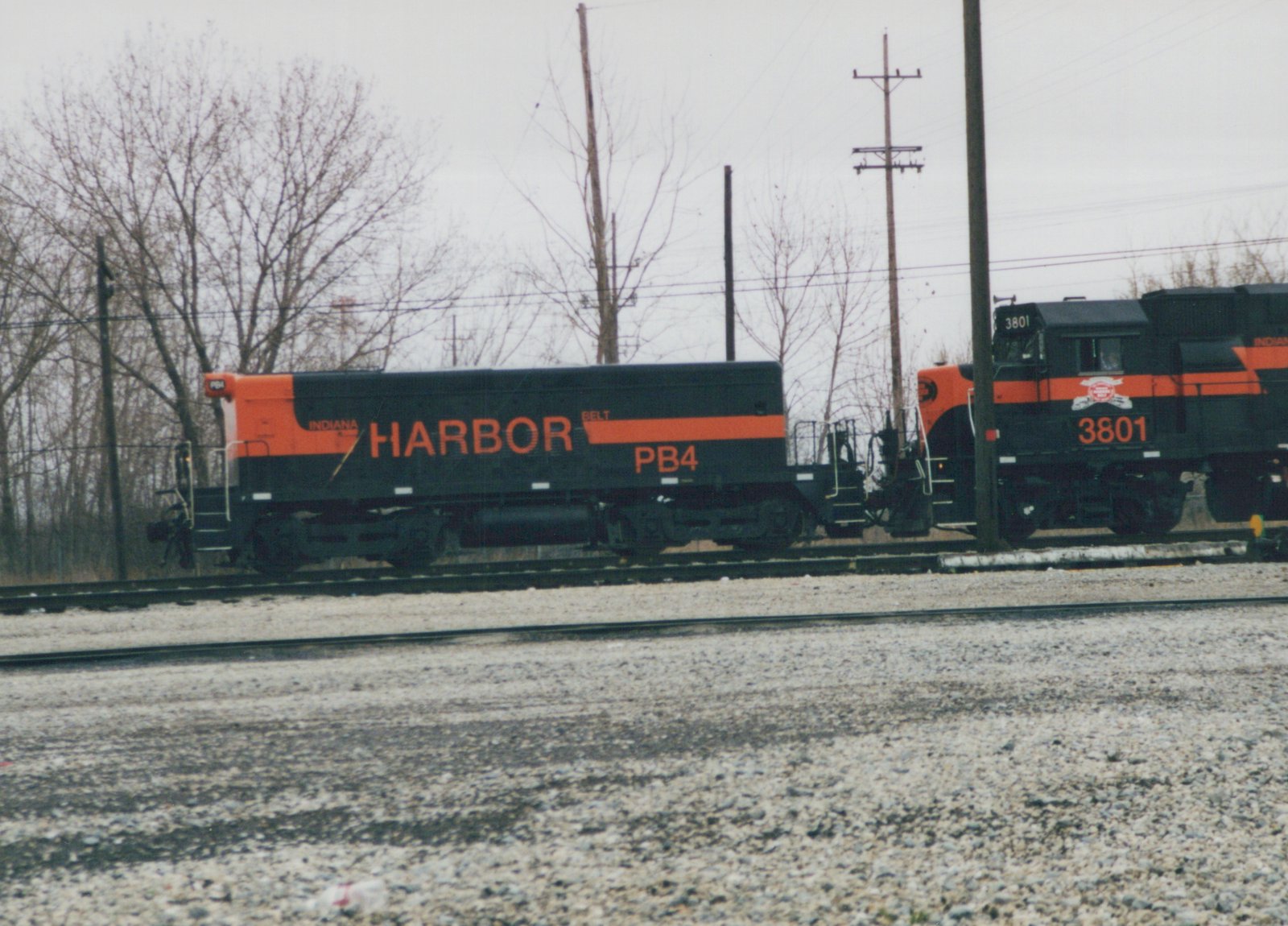 A black and orange cabless unit lettered INDIANA HARBOR BELT PB4, coupled to locomotive 3801, on yard tracks under an overcast sky with bare trees and utility poles behind.
