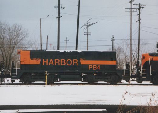 Side view of a black and orange cabless diesel unit lettered INDIANA HARBOR BELT and PB4, standing on a snow-covered track under an overcast sky, with the orange end of a second locomotive at the right.