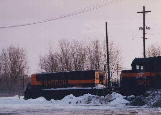 A black and orange cabless locomotive lettered HARBOR and numbered PB4 is coupled to black and orange locomotive 3801. Snowbanks are in front of them and bare trees stand behind under an overcast sky.
