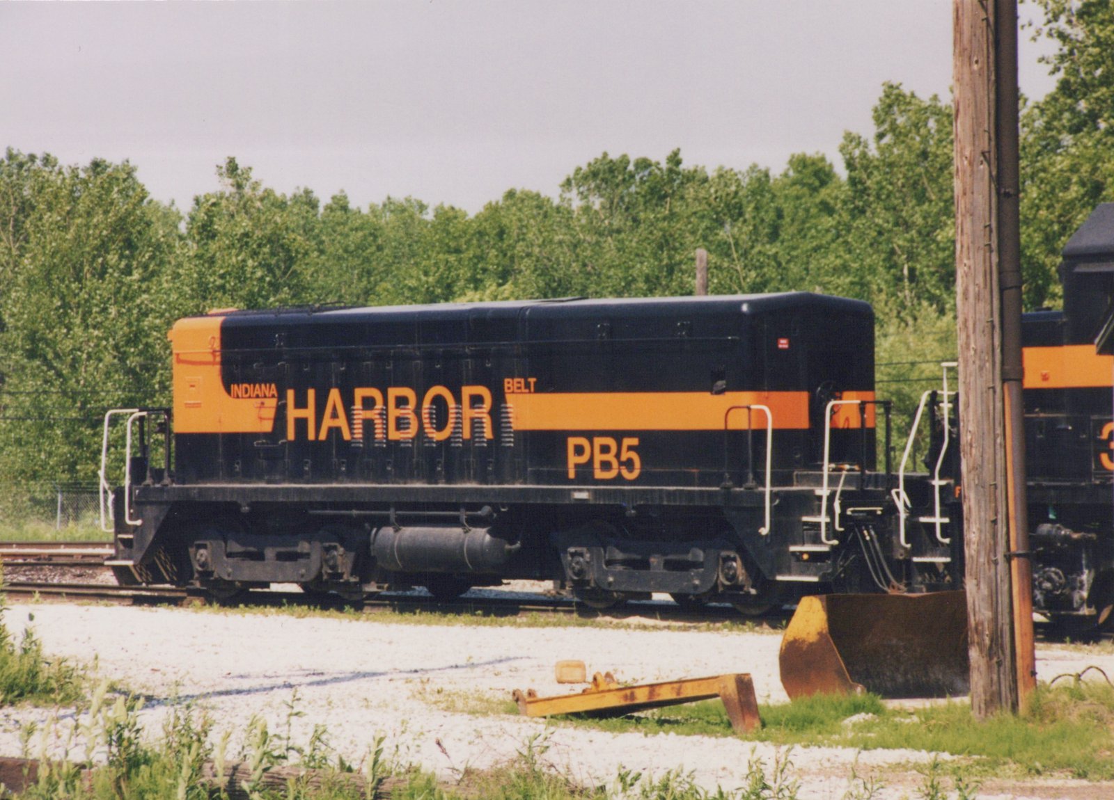Side view of a black cabless locomotive unit with an orange stripe and the lettering INDIANA HARBOR BELT and PB5, parked on a yard track with trees behind it and a rusty bucket and a yellow steel beam on the gravel in front.